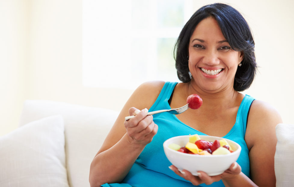 Overweight Woman Sitting On Sofa Eating Bowl Of Fresh Fruit;