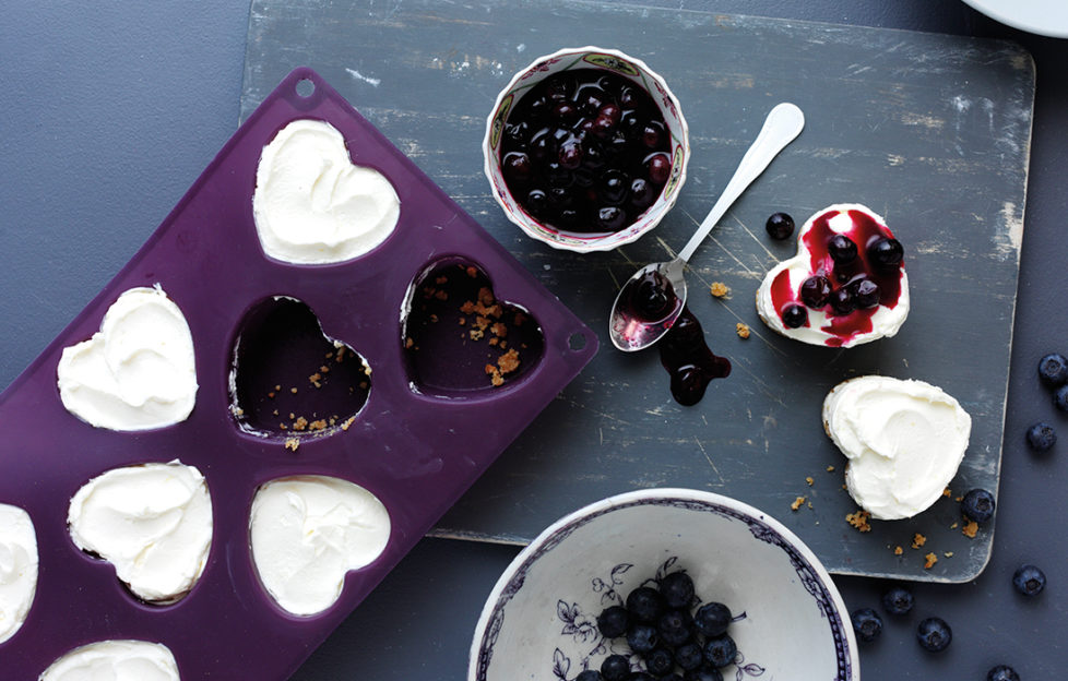 Purple silicone mould with heart shaped holes, all but 2 full of cheesecake mix, 2 cheesecakes removed and topped with blueberries mix