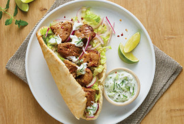 Toasted pitta bread on plate with salad and golden brown chunks of lamb, small pot of yogurt and cucumber on the side