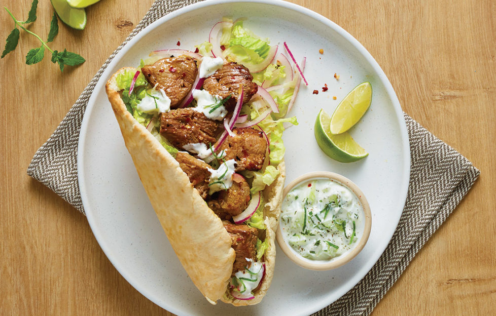 Toasted pitta bread on plate with salad and golden brown chunks of lamb, small pot of yogurt and cucumber on the side