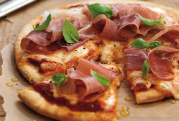Pizza decorated with ham and basil leaves, one slice cut