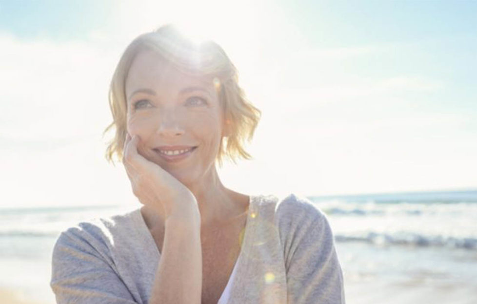 Smiling woman on beach, wearing sleeved top, sun behind
