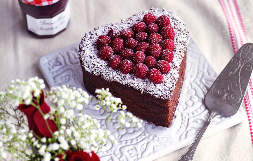 Heart shaped chocolate Valentine cake topped with fresh raspberries, on table with vase of red roses and gypsophila and jar of Bonne Maman conserve