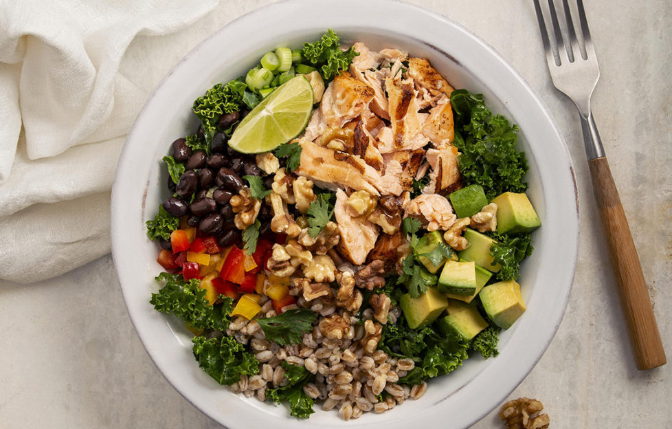 Bowl of salad with flaked salmon, crushed walnuts, cubed pepper and avocado, kale and lime wedge