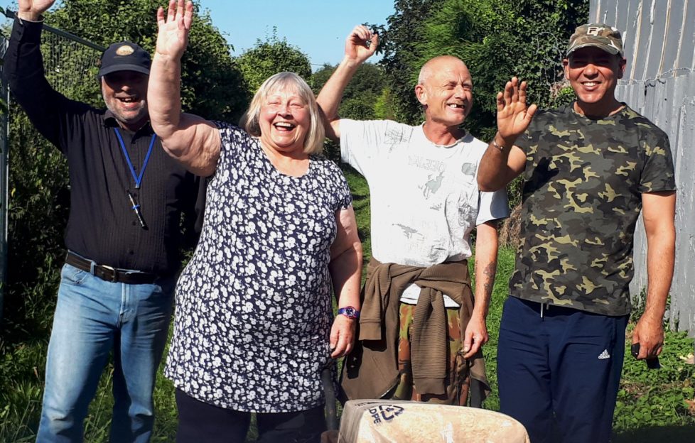 Four people at allotments with wheelbarrow, smiling and waving
