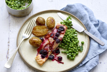 Plate with salmon fillet topped with cooked blueberries and drizzle of deep red sauce, roast potatoes and peas on the side
