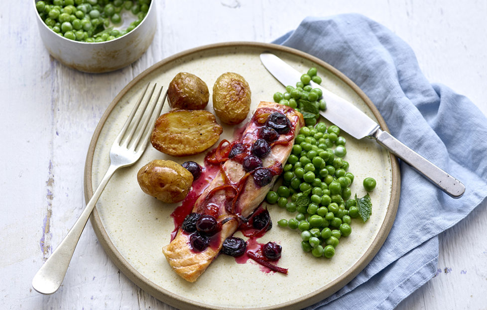 Plate with salmon fillet topped with cooked blueberries and drizzle of deep red sauce, roast potatoes and peas on the side