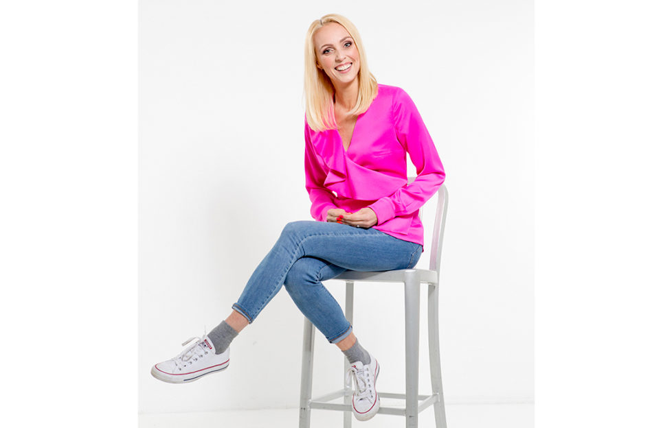 Happy slim blonde woman in bright pink top and blue jeans posing on stool, white background