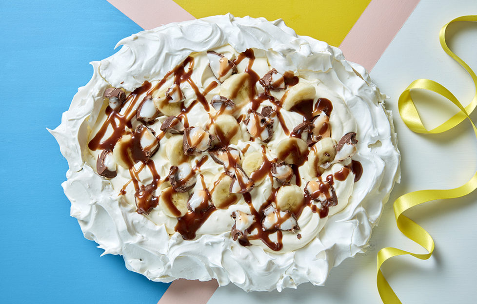 Pavlova with oval egg white edge, and creamy filling topped with banana slices, chopped Creme Egg pieces and chocolate sauce drizzle
