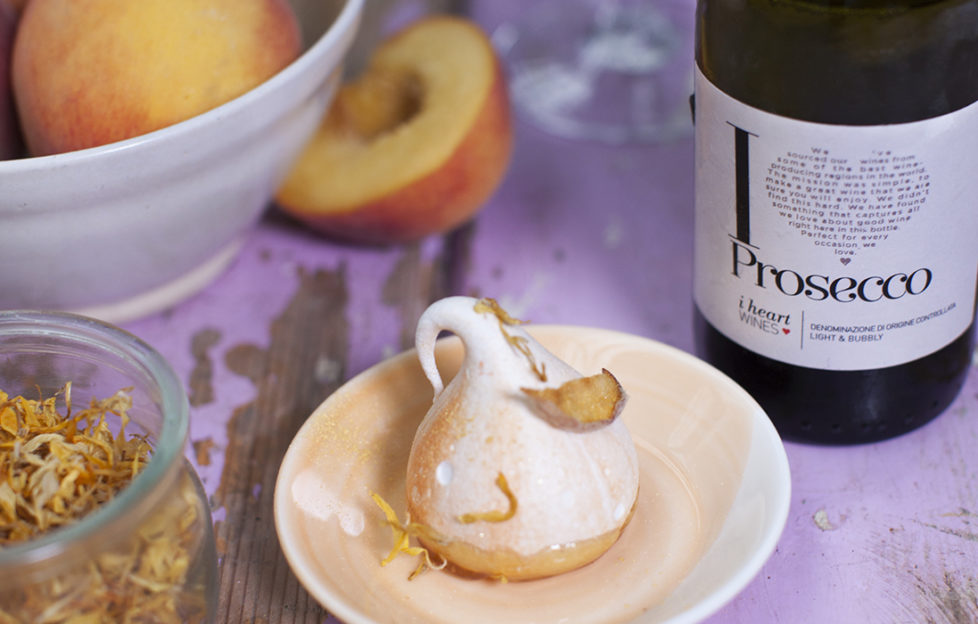 Meringue decorated with a petal and gold swirls, bowl of peaches and bottle of i heart prosecco