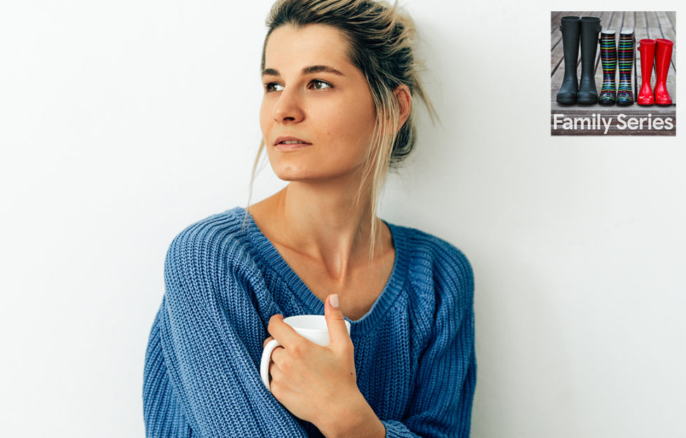 Pensive woman alone on Mother's Day in baggy blue jumper holds white mug and gazes into the distance