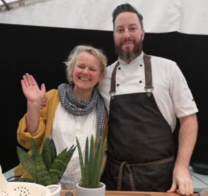 Mature woman smiling and waving with young bearded chef, in marquee