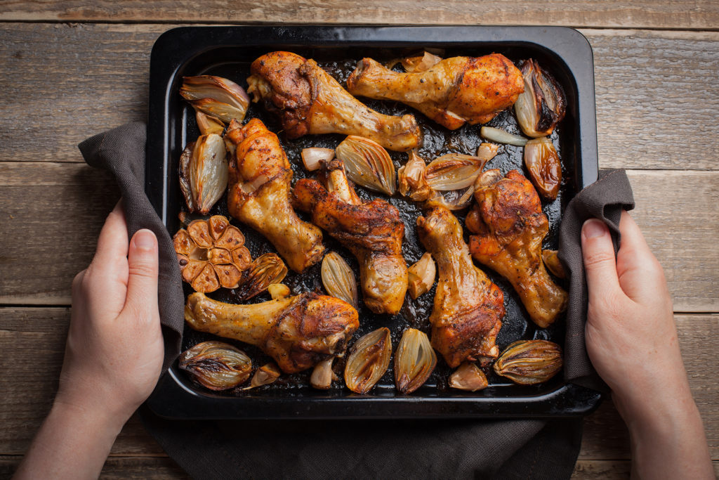 Female hands holding hot baking sheet from oven baked chicken legs with shallots and garlic. A woman puts on a dark wooden table with a tray of fried chicken. 