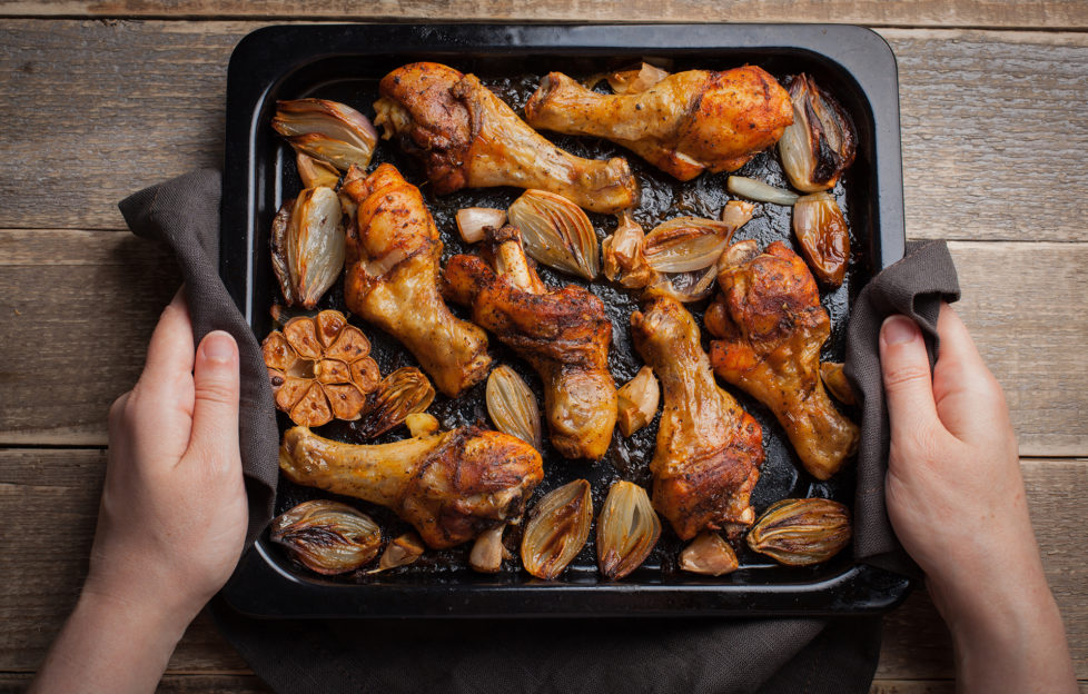 Female hands holding hot baking sheet from oven baked chicken legs with shallots and garlic. A woman puts on a dark wooden table with a tray of fried chicken.