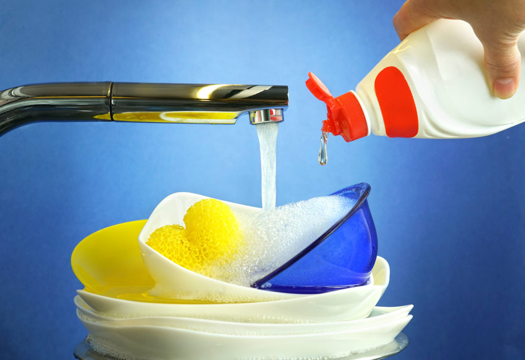 Concept washing dishes in kitchen on blue background close-up. Stream of water from tap and hand holds bottle with liquid detergent. Dish washing liquid drops fall dripping