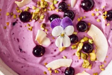 Close up of blackcurrant smoothie bowl ttopped with seeds, fresh blackcurrants and a small purple pansy flower