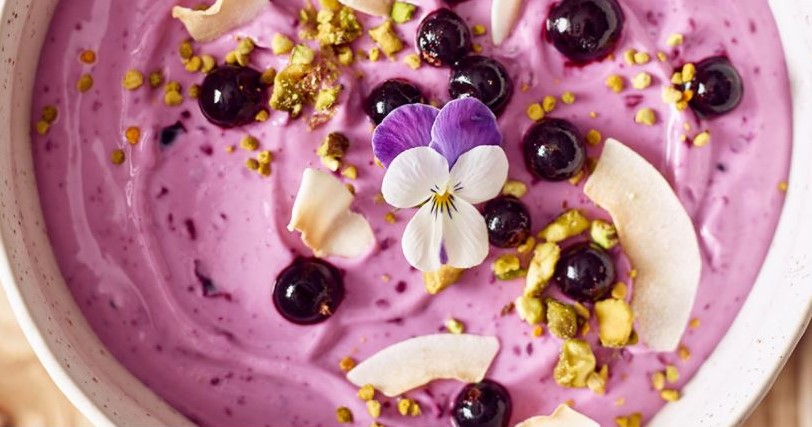 Close up of blackcurrant smoothie bowl ttopped with seeds, fresh blackcurrants and a small purple pansy flower