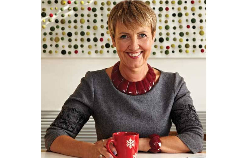 Aggie Mackenzie, stylish mature woman with short blonde hair in grey top with burgundy statement necklace, sitting smiling in kitchen with red spotted mug