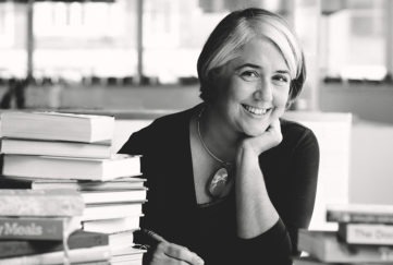 Black and white image, Alison Oakervee, stylish mature woman with part-blonde bob hairstyle, smiling and relaxed, at desk with pile of cookery books. Advice on how to freeze food