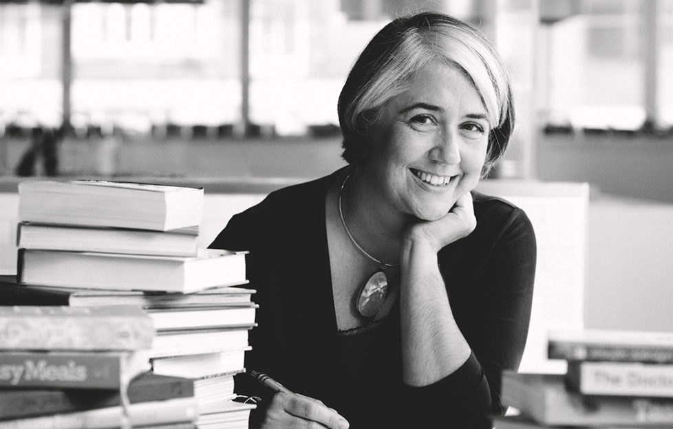 Black and white image, Alison Oakervee, stylish mature woman with part-blonde bob hairstyle, smiling and relaxed, at desk with pile of cookery books. Advice on how to freeze food