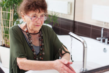 Bohemian elderly woman in loose layered tops and jewellery washing hands under kitchen tap