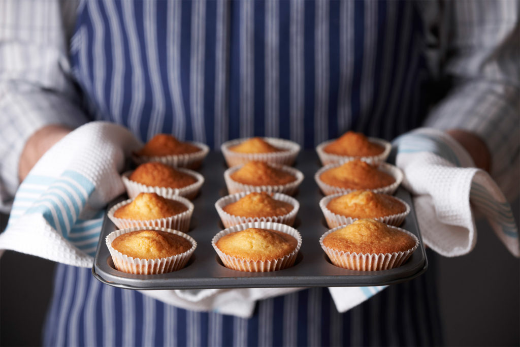 Person in striped apron holding tray of 12 freshly baked cupcakes with a tea towel