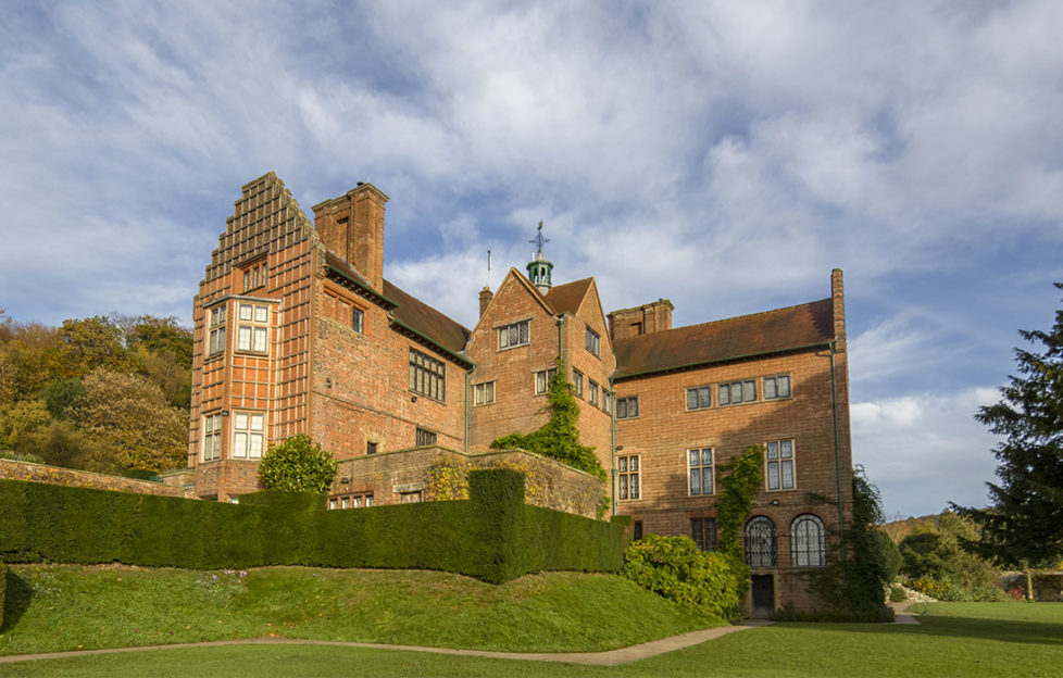 Red brick mansion house with row of arched windows, stepped gable, square chimney stacks and neatly clipped hedge, blue sky behind