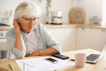 Mature woman at kitchen table sorting out finances with paper statements, phone, calculator and laptop, also mug of tea