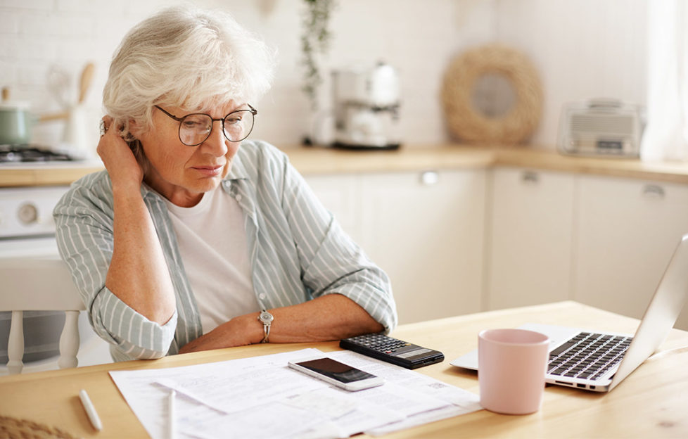 Mature woman at kitchen table sorting out finances with paper statements, phone, calculator and laptop, also mug of tea