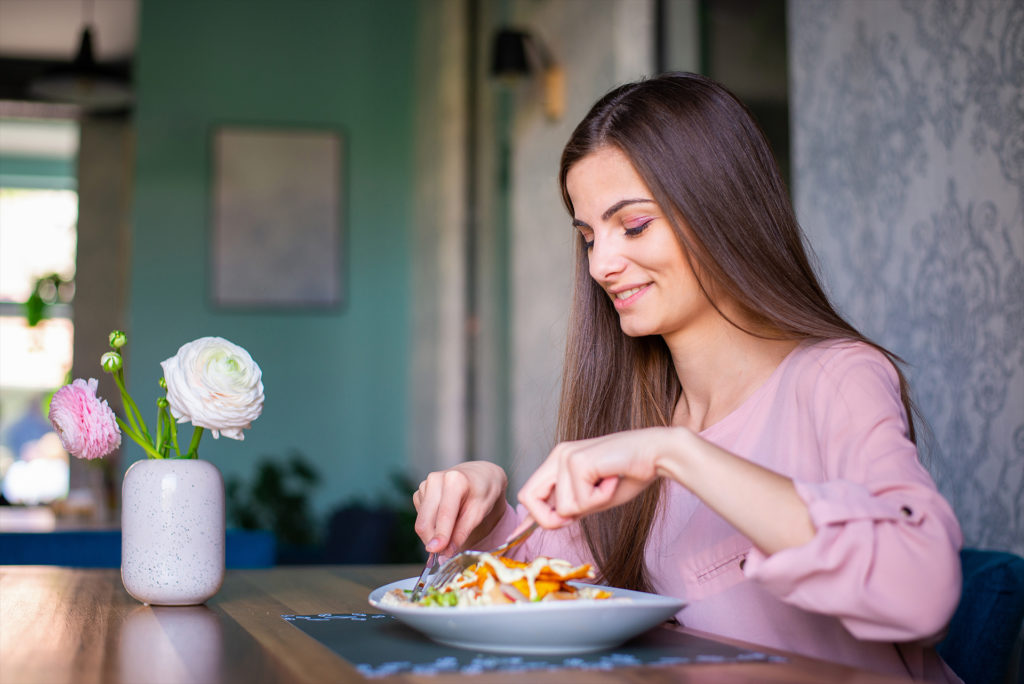 Pretty young woman with long hair and in pink shirt eating a salad at the nice restaurant. Eating alone. Comfortable, happy, smiling, carefree.