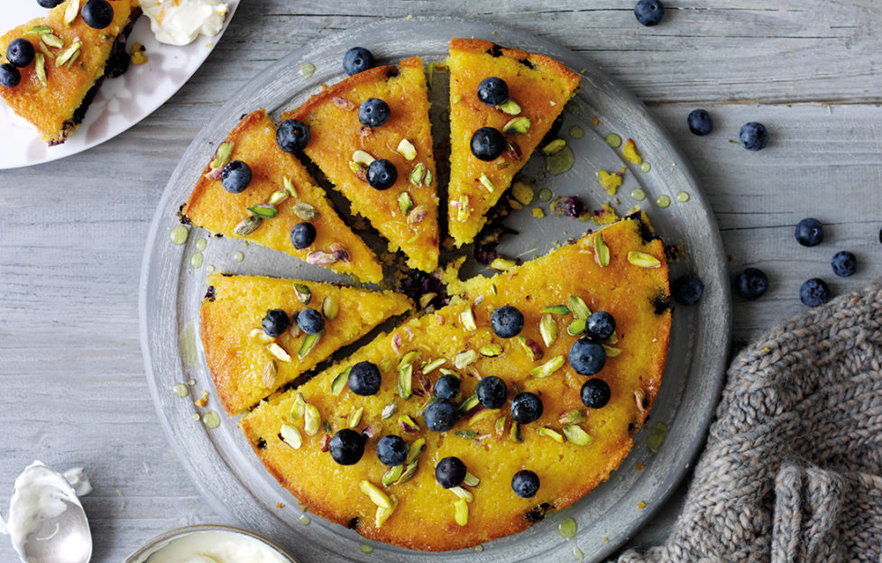 Moist golden spongy cake topped with blueberries and pistachios, 5 slices cut, 1 on a plate