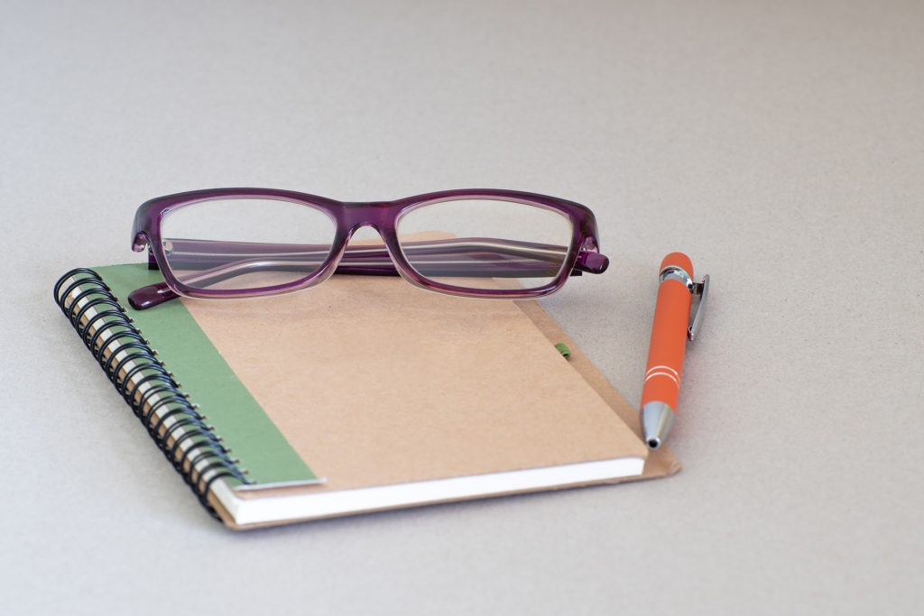 Recycled blank brown front cover spiral notebook with orange pen and purple reading glasses isolated on background. Adult night class note taking. Female/woman/mother's notes. 
