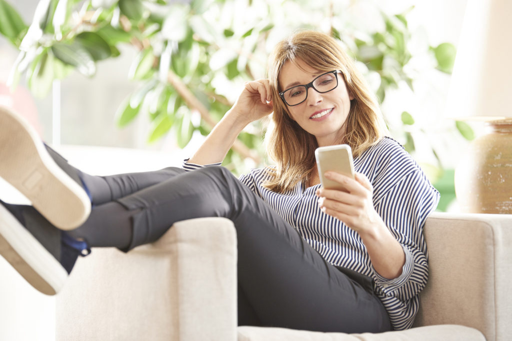 Full length shot of a smiling mature woman reading message on her mobile phone while sitting on armchair at home.; 
