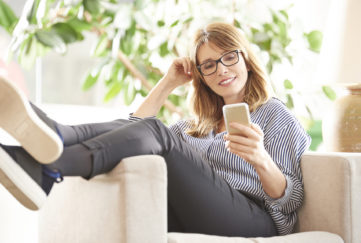 Full length shot of a smiling mature woman reading message on her mobile phone while sitting on armchair at home.;