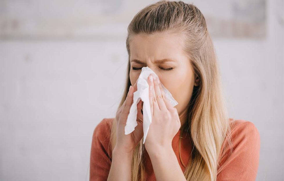 blonde woman sneezing while holding tissue near nose