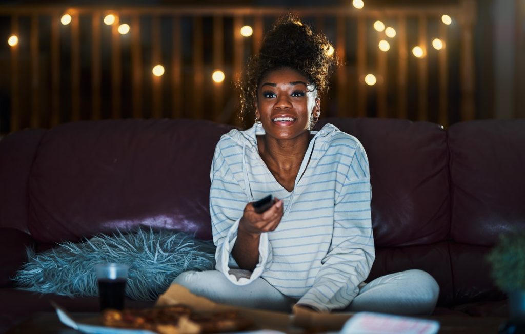 Woman smiling as she aims remote control at TV, night time city lights visible through window behind her