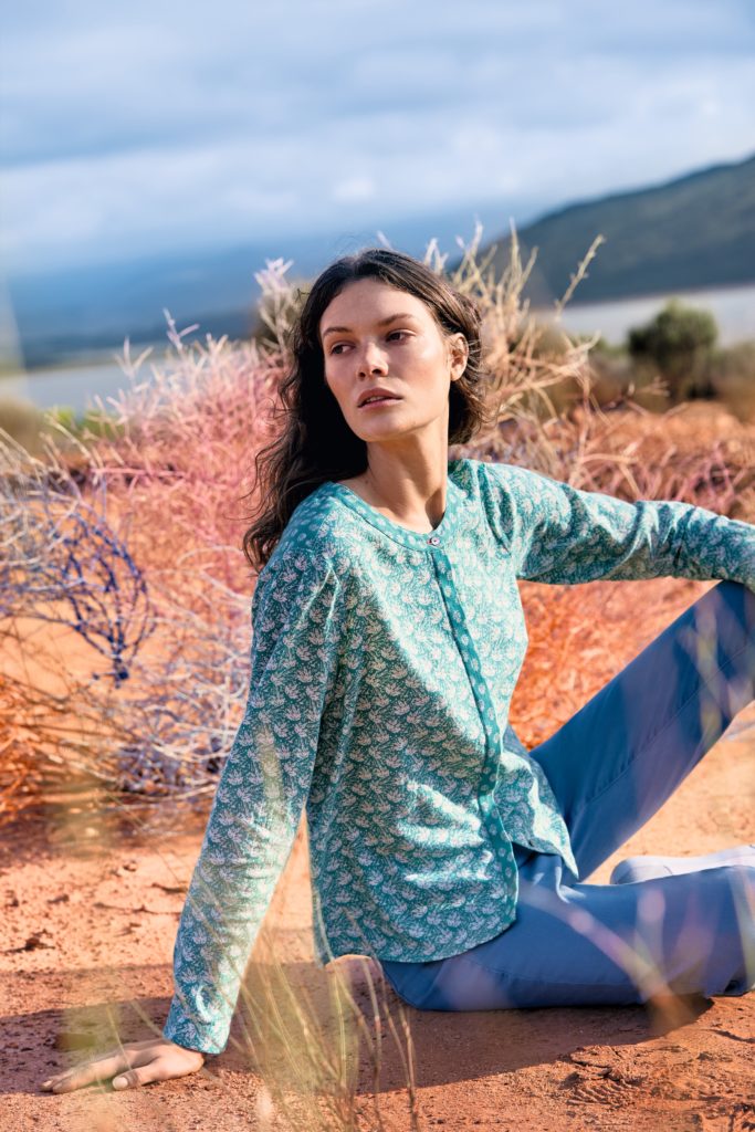 Model sitting in arid landscape, wearing blue jeans and floaty turquoise shirt, round neck, no collar, with small repeating white pattern