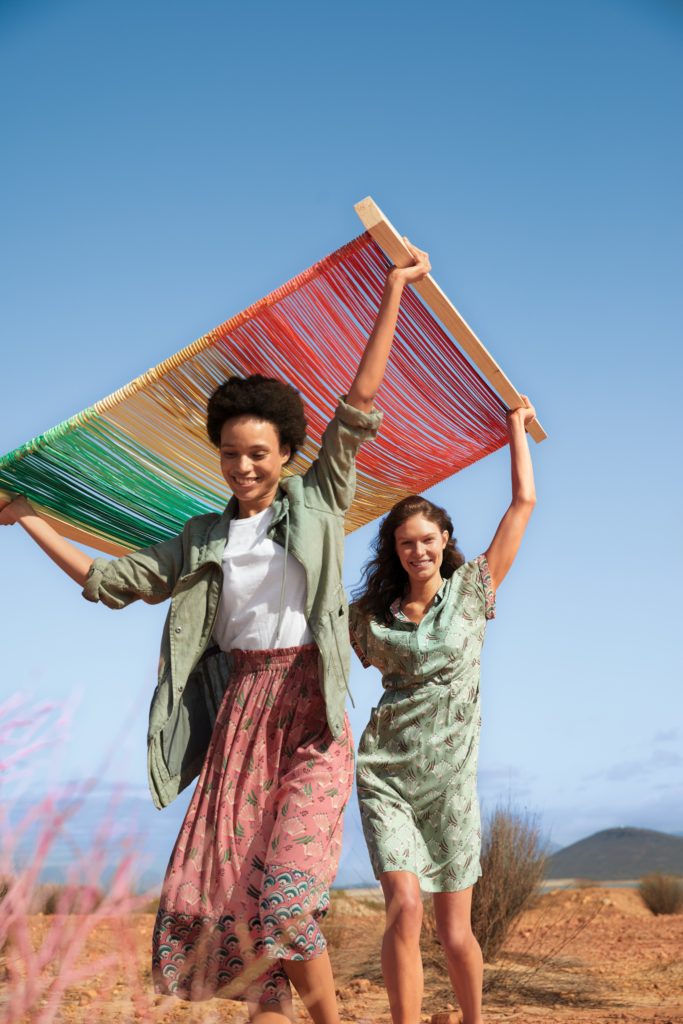 Two models carry a loom with coloured yarn strands. One wears a mint green cotton dress and the other a long casual patterned pink skirt, white t shirt and long shirt-style khaki jacket
