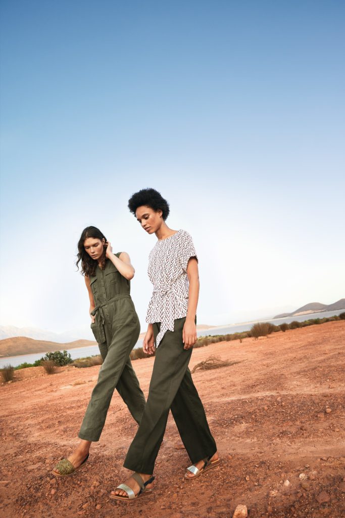 2 models walking on arid ground, one wears sleeveless khaki jumpsuit, the other straight leg, olive green trousers and a relaxed casual fit patterned blouse with tie belt 