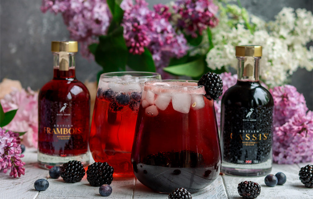 Two bulbous glasses of deep red cocktail drink, 2 bottles, one of cassis and one of framboise, with a vase of lilac blooms behind