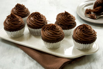 Six chocolate cupcakes on a white rectangular plate, all with large perfect swirls of chocolate fudge icing