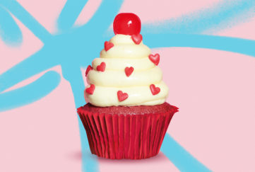 Red velvet cupcake with tower of white buttercream studded with tiny red sugar hearts and topped with a cherry, pink/blue background.