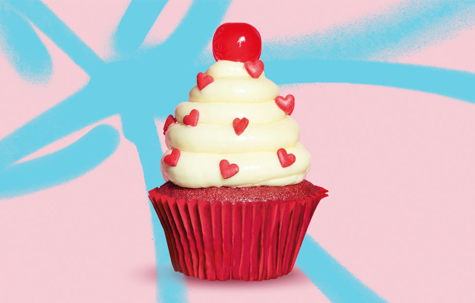 Red velvet cupcake with tower of white buttercream studded with tiny red sugar hearts and topped with a cherry, pink/blue background.