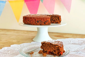 Rich brown fruit cake on a cake stand, one slice cut on a plate, bunting hung behind