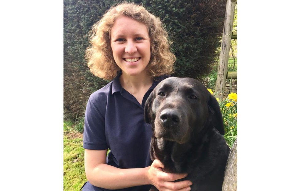 Smiling young woman with wavy blonde hair, in dark polo shirt, in garden hugging black Labrador