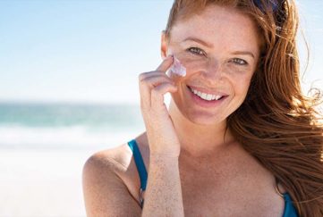 Smiling young woman applying sunscreen lotion on face at beach, with copy space. Portrait of beautiful happy girl using sunblock on her delicate skin with freckles and looking at camera.
