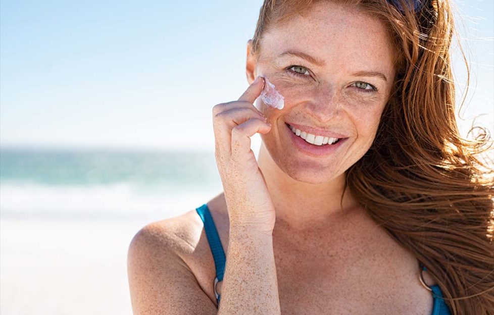 Smiling young woman applying sunscreen lotion on face at beach, with copy space. Portrait of beautiful happy girl using sunblock on her delicate skin with freckles and looking at camera.