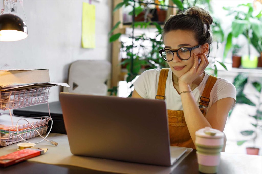 Young female gardener in glasses using laptop, communicates on internet with customer in home garden/greenhouse, reusable coffee/tea mug on table.Cozy office workplace, remote work, E learning concept