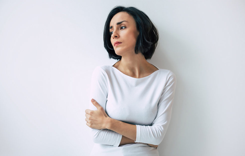 Woman in an anxious mood, arms wrapped around herself, looking to the side with sad expression, against plain white wall