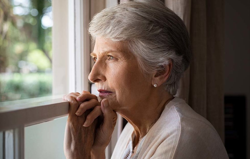 Depressed senior woman at home feeling sad. Elderly woman looks sadly outside the window. Depressed lonely lady standing alone and looking through the window.